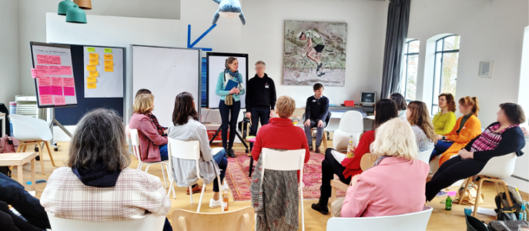 Personen in einem Stuhlhalbkreis Workshop-Raum. Vorne stehen zwei moderierende Personen.