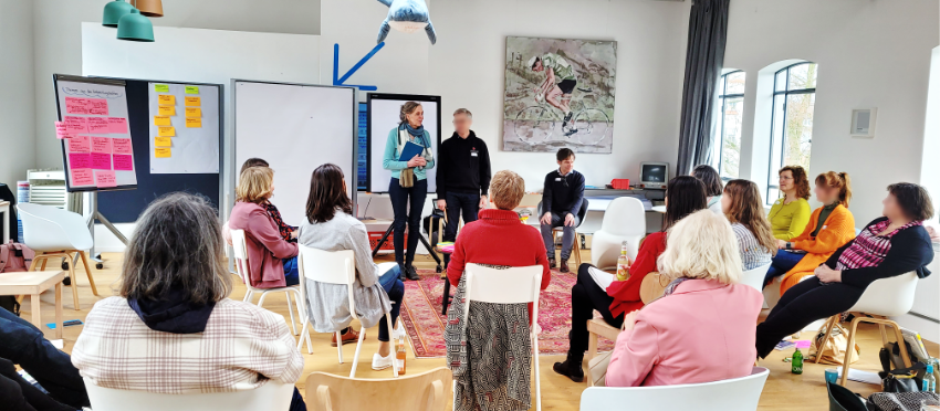 Personen in einem Stuhlhalbkreis Workshop-Raum. Vorne stehen zwei moderierende Personen.